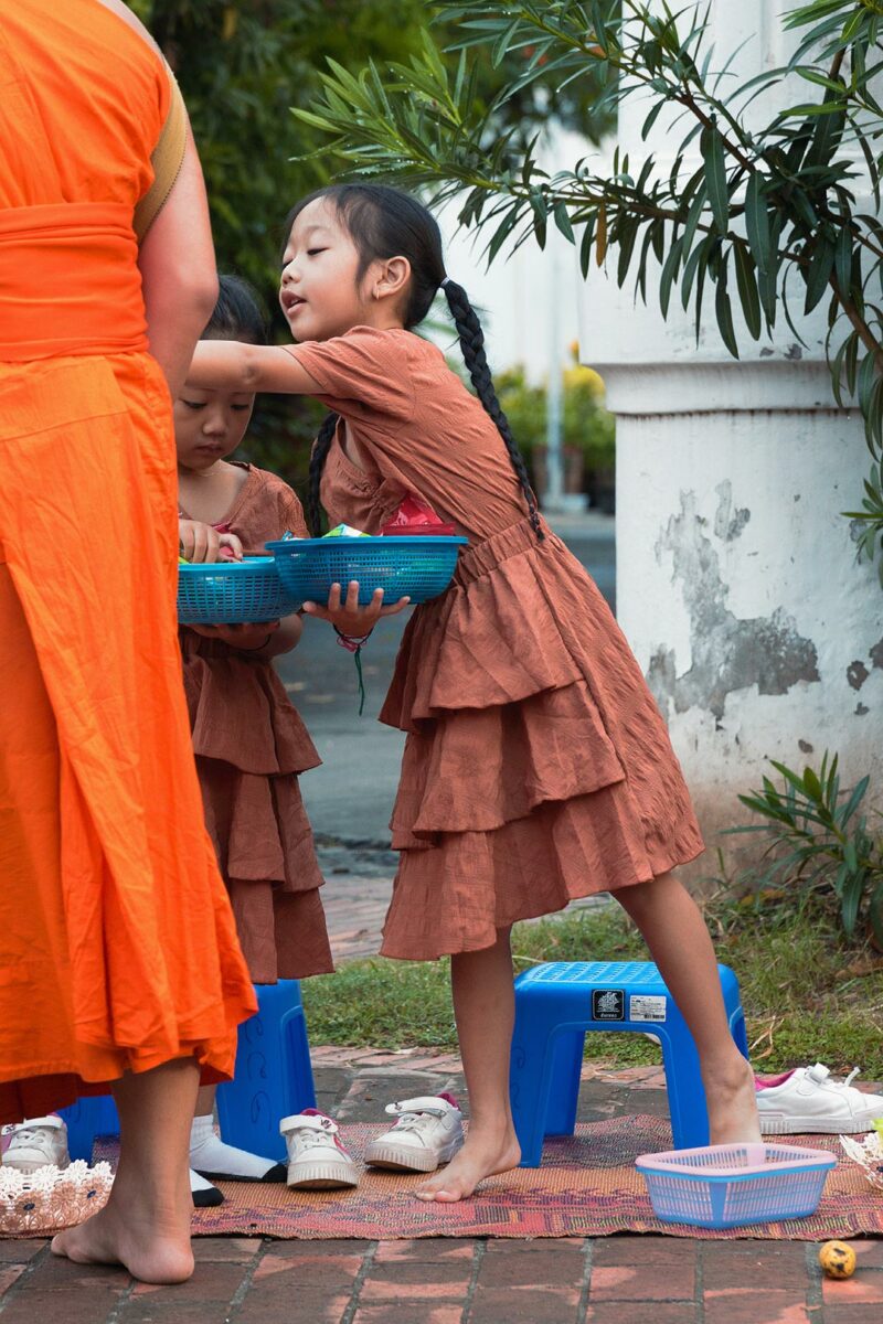 Little asian girl giving food to a young monk at the Alms Ceremony of Luang Prabang in Laos